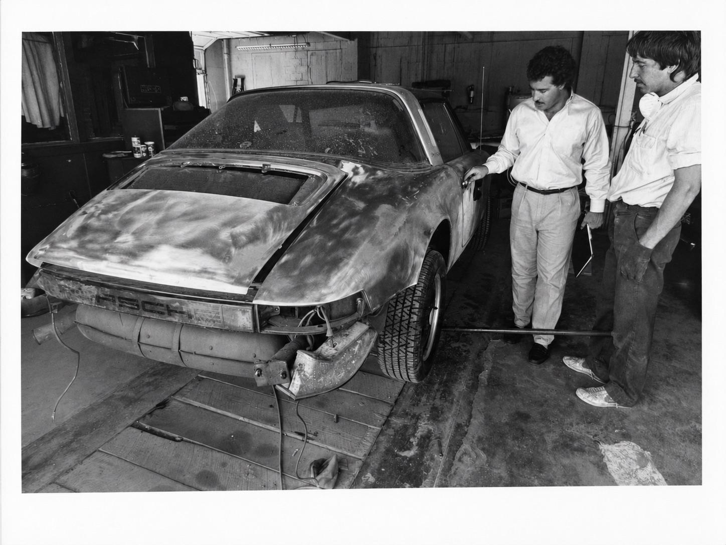 Black-and-white archival photograph of two young men in a body shop inspecting the rear-end damage on a stripped Porsche 924