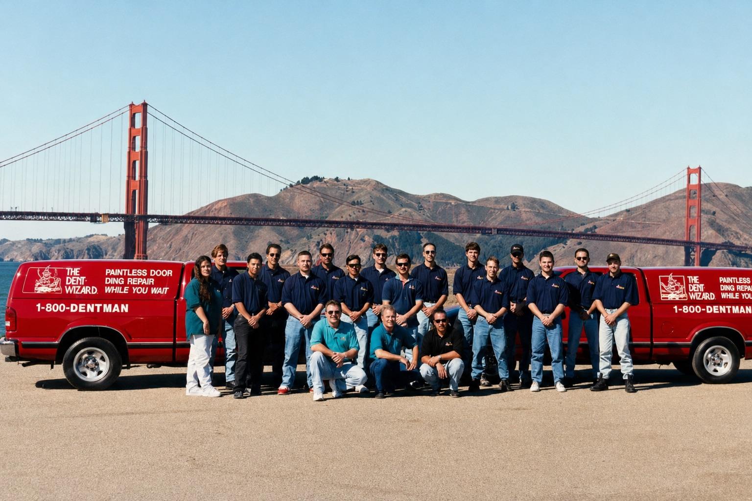 Color group photograph of approximately twenty Dent Wizard technicians standing in front of two red 1-800-DENTMAN service vans with the Golden Gate Bridge in the background